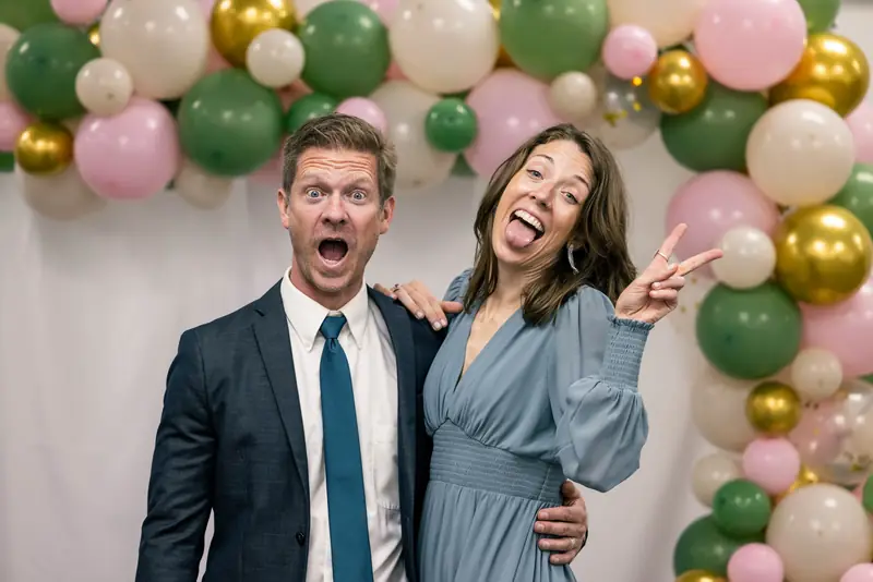 A couple making silly faces in front of a balloon arch